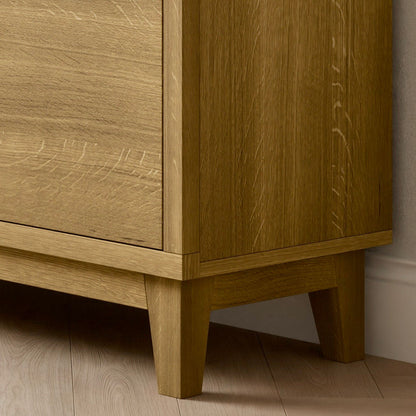 A close-up of the Double Drawer, Double Door Media Cabinet in White Oak by Krovel Furniture Co. shows its handmade design, resting on a light wood floor. The cabinet features a smooth brown finish with visible grain patterns and stands on four short, angled legs.
