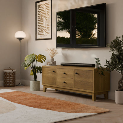 A modern living room with the Krovel Furniture Co. Maine Made Double Drawer, Double Door Media Cabinet, a wall-mounted TV, potted plants, floor lamp, abstract art piece, and a beige-and-orange area rug on light wooden flooring.