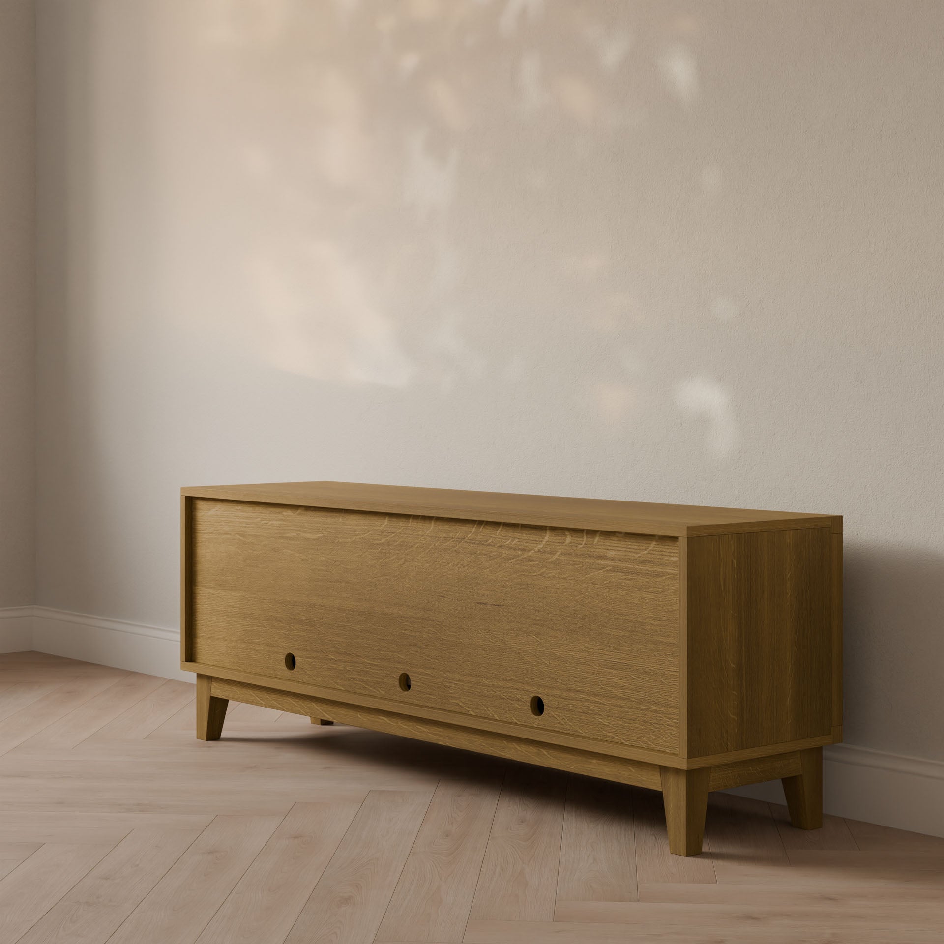 The Maine Made Double Drawer, Double Door Media Cabinet by Krovel Furniture Co. sits against a light-colored wall on wooden flooring, while soft natural light creates a dappled effect on the wall above.