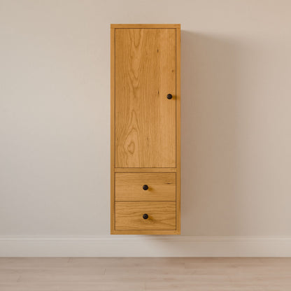 The Maine Made Double Drawer Linen Cabinet by Krovel Furniture Co. features a solid hardwood build with a vertical door above two drawers, finished with round black knobs, and is set against a light-colored wall and floor.