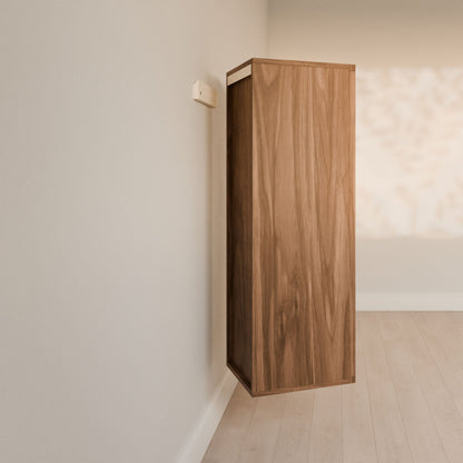 A wall-mounted Double Drawer Linen Cabinet in Walnut by Krovel Furniture Co. is set in a minimalist room with light wood flooring. A nearby small shelf matches the decor, while soft light accentuates its sustainable craftsmanship.