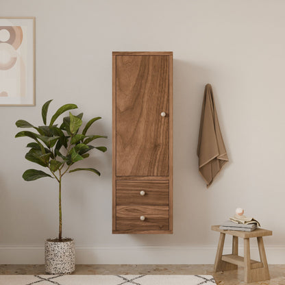 A minimalist room showcases a Krovel Furniture Co. Double Drawer Linen Cabinet in Walnut, wall-mounted in customizable sizes. Below, a small stool with a candle and jar rests on the floor. A potted plant and abstract art decorate the beige wall, near a hanging beige towel.