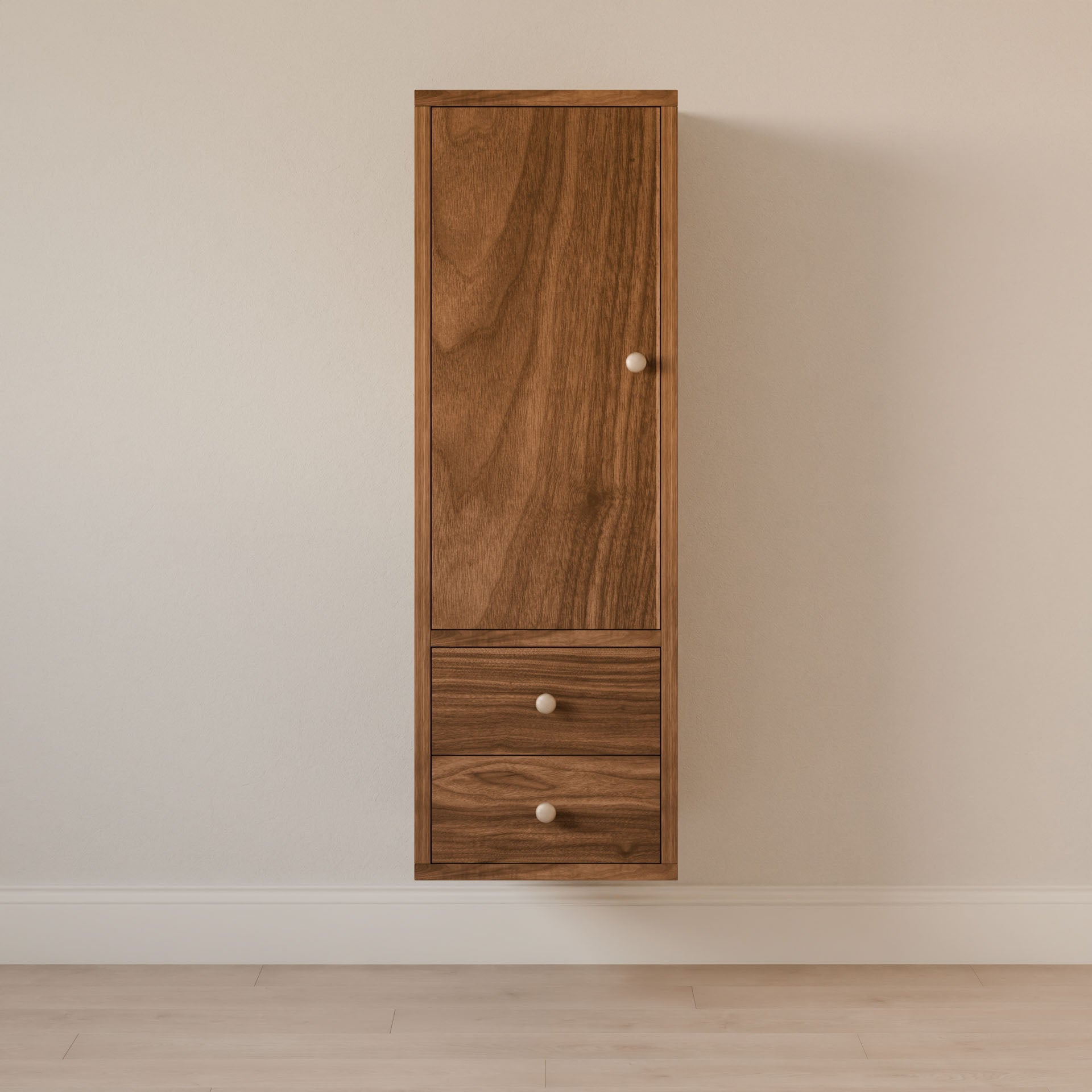 A tall, wall-mounted Krovel Furniture Co. Double Drawer Linen Cabinet in Walnut stands against a plain, light-colored wall. Crafted sustainably, it has a top door and two lower drawers with small round handles. The floor is light wood.
