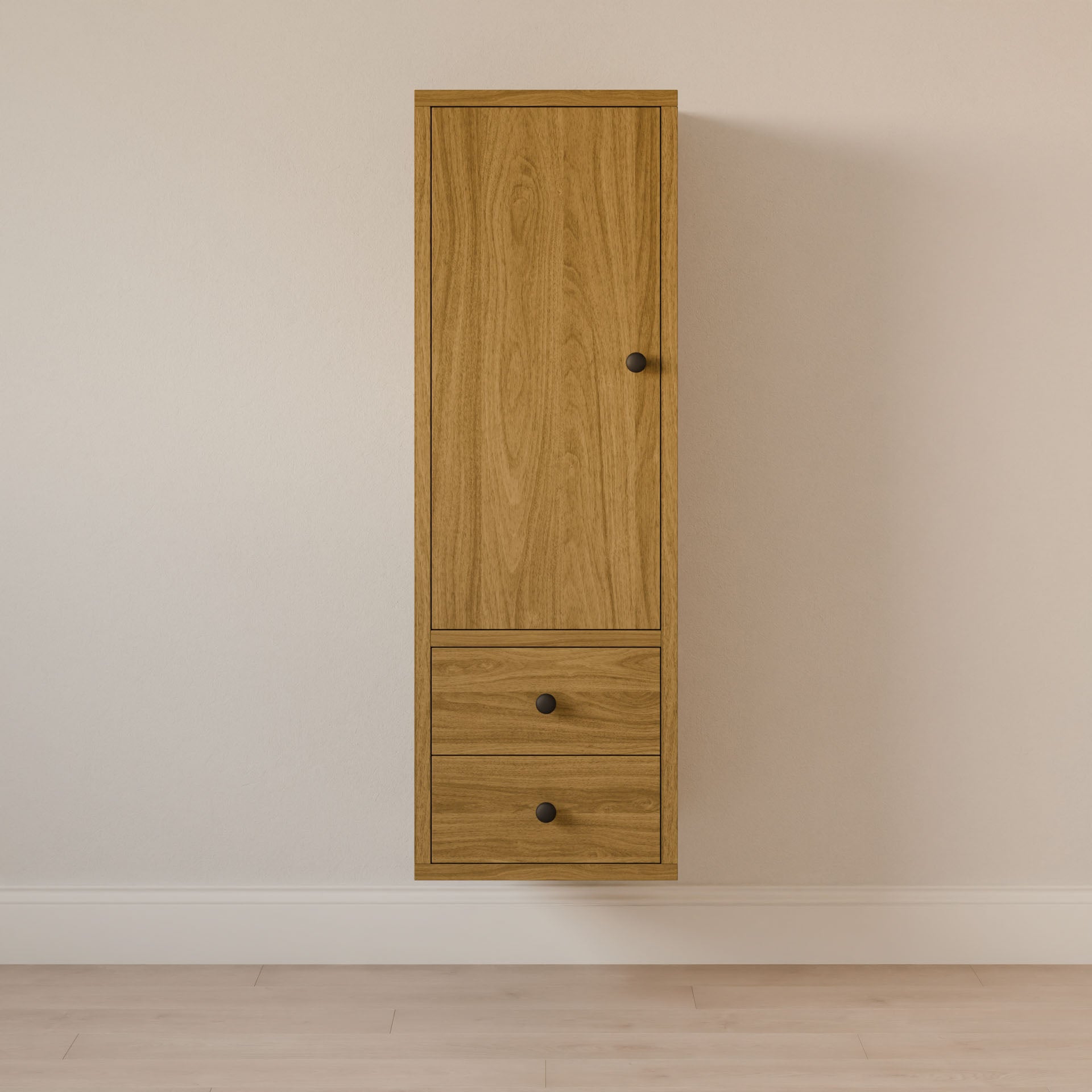 The Double Drawer Linen Cabinet in White Oak by Krovel Furniture Co. is wall-mounted with a tall door and two drawers with black knobs, crafted sustainably. It rests against a light wall above hardwood flooring and comes in customizable sizes.