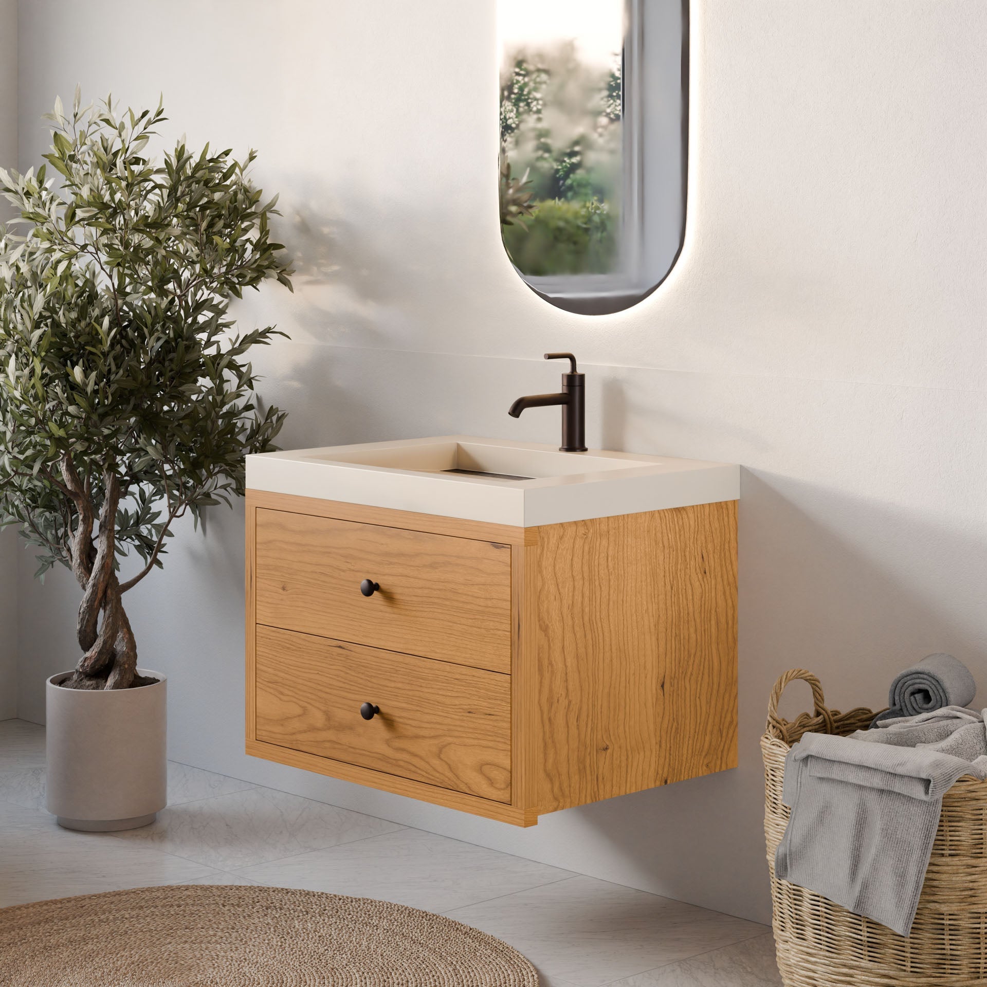 A modern bathroom corner showcases the Krovel Furniture Co. Double Drawer Vanity in Cherry with two custom-size drawers and an oval mirror above. Nearby, a small potted olive tree stands next to a wicker basket with rolled towels on a light-colored floor.