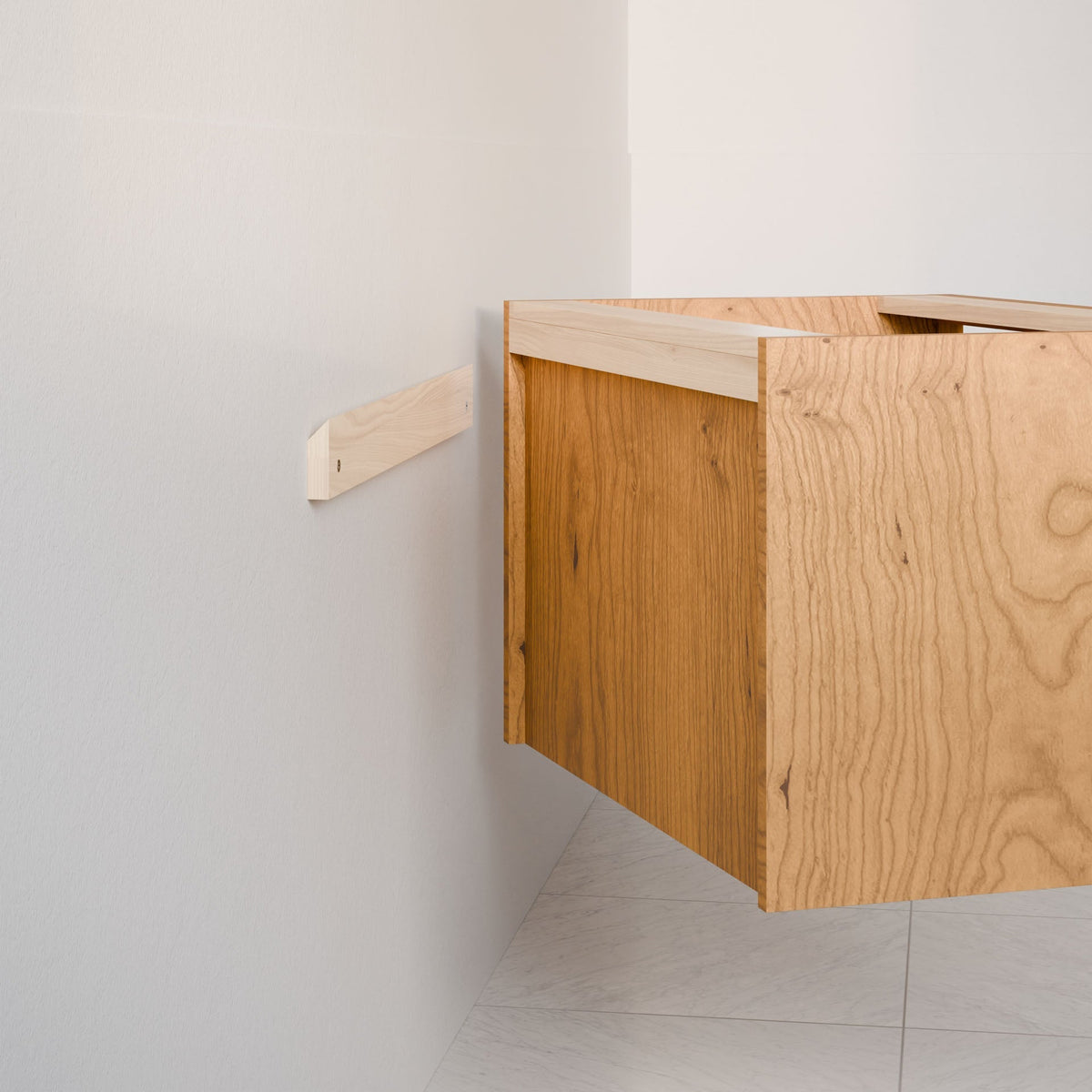 The Double Drawer Vanity in Cherry by Krovel Furniture Co. is partially installed on a white wall with a horizontal cleat. The unfinished cabinet showcases visible wood grain, and the floor features white tiles.