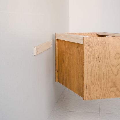 The Double Drawer Vanity in Cherry by Krovel Furniture Co. is partially installed on a white wall with a horizontal cleat. The unfinished cabinet showcases visible wood grain, and the floor features white tiles.