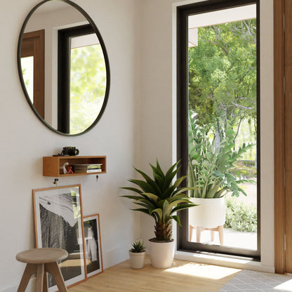 A modern, light-filled entryway features a round mirror and Krovel's Entryway Organizer in Cherry, with two plants. Artwork leans casually against the wall, while a large potted plant near the glass door offers views of the lush garden beyond.