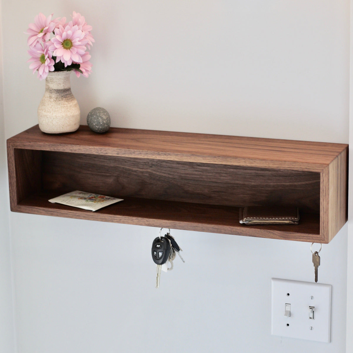 The Krovel Entryway Organizer in Walnut holds a vase with pink flowers, a small stone, and mail. Keys and a keychain hang from hooks below this handmade key holder, which is mounted above a light switch on a light wall.