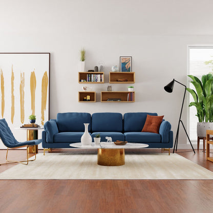 A modern living room showcases a blue sofa, white rug, and a marble coffee table with a gold base. A blue chair sits to the left beneath Krovel's Floating Bookshelf in Maple displaying decor. Handmade wall shelves add charm alongside an abstract painting, floor lamp, and plants.