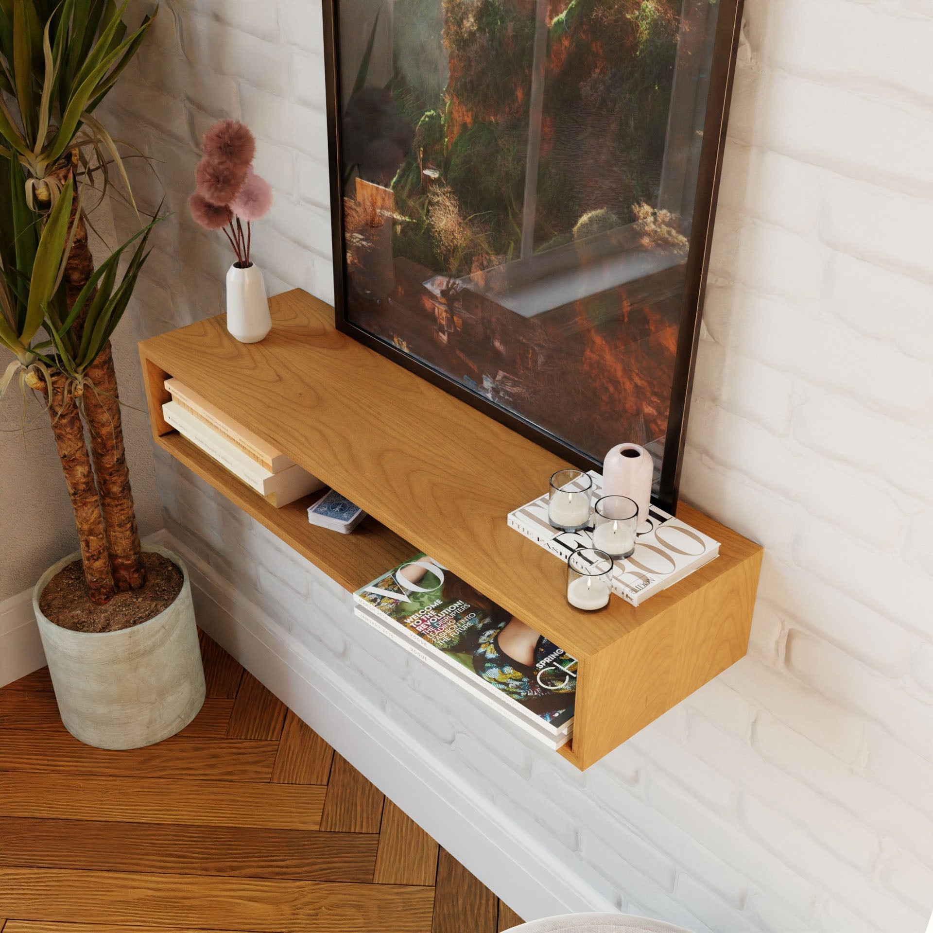A Krovel Floating Console Table in Cherry enhances a white brick wall, displaying pottery, candles, books, and a magazine. A large framed picture rests on the console. Nearby, a tall plant in a gray pot matches the wooden floor for a mid-century modern vibe.