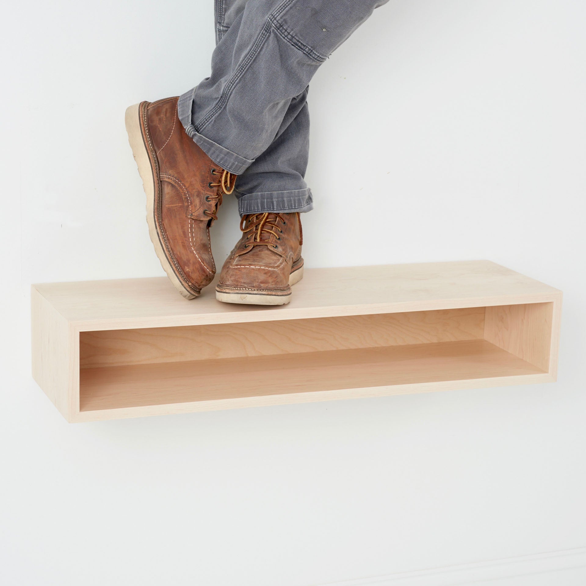 A person in brown boots and gray pants stands atop a Krovel Hardwood Floating Console Table mounted on a white wall.