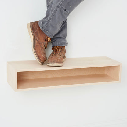 A person in brown boots and gray pants stands atop a Krovel Hardwood Floating Console Table mounted on a white wall.