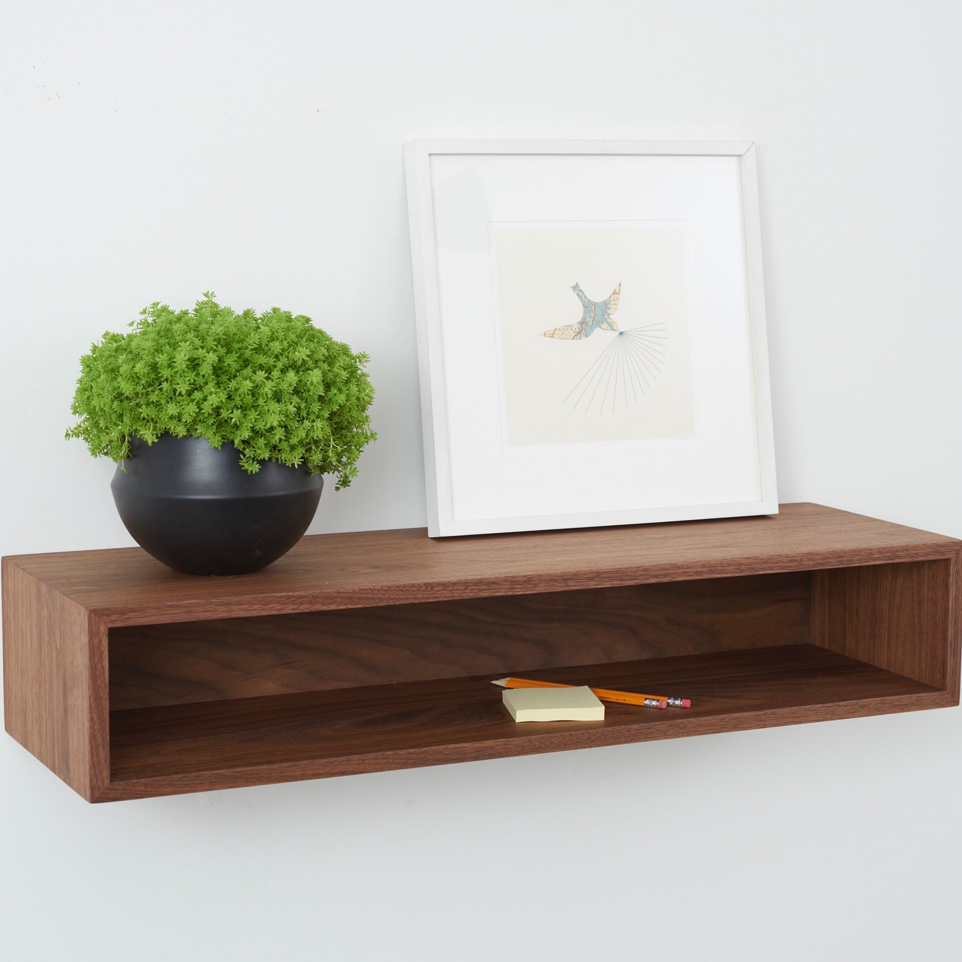 The Krovel Hardwood Floating Console Table displays a small black pot with a green plant, a framed art print, a pencil, and two sticky note pads against a plain white wall.