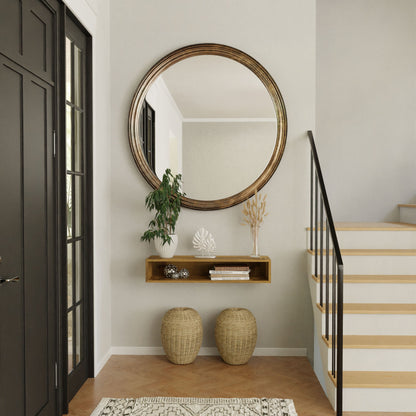 A modern entryway showcases the Krovel Hardwood Floating Console Table under a large round mirror, adorned with plants, a vase, and books. Two woven baskets sit below the shelf beside a wooden staircase.