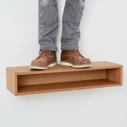 A person wearing gray pants and brown boots stands on a Krovel Hardwood Floating Console Table mounted on a white wall.