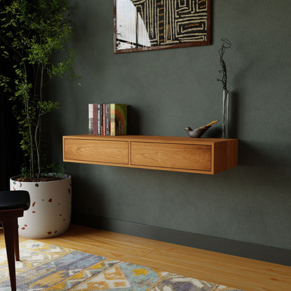A modern living room features Krovel's Floating Desk in Cherry, adorned with books and a vase. A decorative bowl adds charm to the display against a dark green wall with abstract art. A large potted plant and part of a rug enhance the wooden floor.