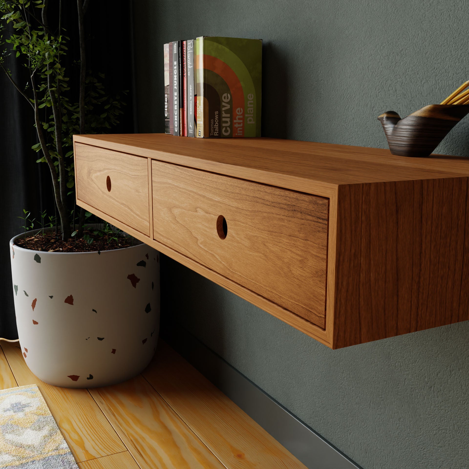 On a green wall, the Krovel Floating Desk in Cherry, featuring two drawers, showcases books and a small pot with pencils. Beside it stands a potted plant, enhancing the modern minimalist ambiance. A rug is partially visible on the wooden floor.