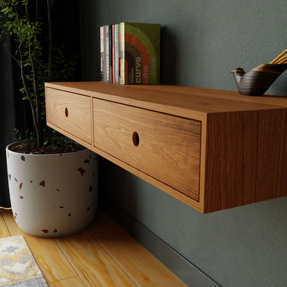 On a green wall, the Krovel Floating Desk in Cherry, featuring two drawers, showcases books and a small pot with pencils. Beside it stands a potted plant, enhancing the modern minimalist ambiance. A rug is partially visible on the wooden floor.