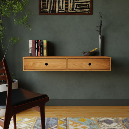 A minimalist room features a Krovel Floating Desk in Cherry, displaying books and a small sculpture. Beneath, a patterned rug adorns the wooden floor. A chair is partially visible on the left, with a plant standing in the corner against a textured green wall.