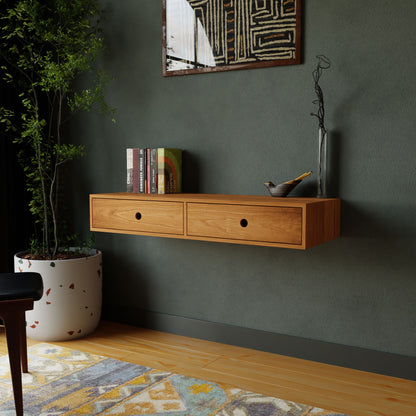 A modern room showcases a Krovel Floating Desk in Cherry with two drawers. On top, books and a vase sit elegantly. The dark green wall features abstract art, complemented by a nearby large potted plant. A colorful rug on the wooden floor highlights handmade details throughout.