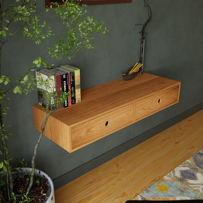 A handmade Krovel Floating Desk in Cherry with two drawers is mounted on a wall beside a leafy plant. The desk holds books and a pencil holder, above wooden flooring and alongside a patterned rug.