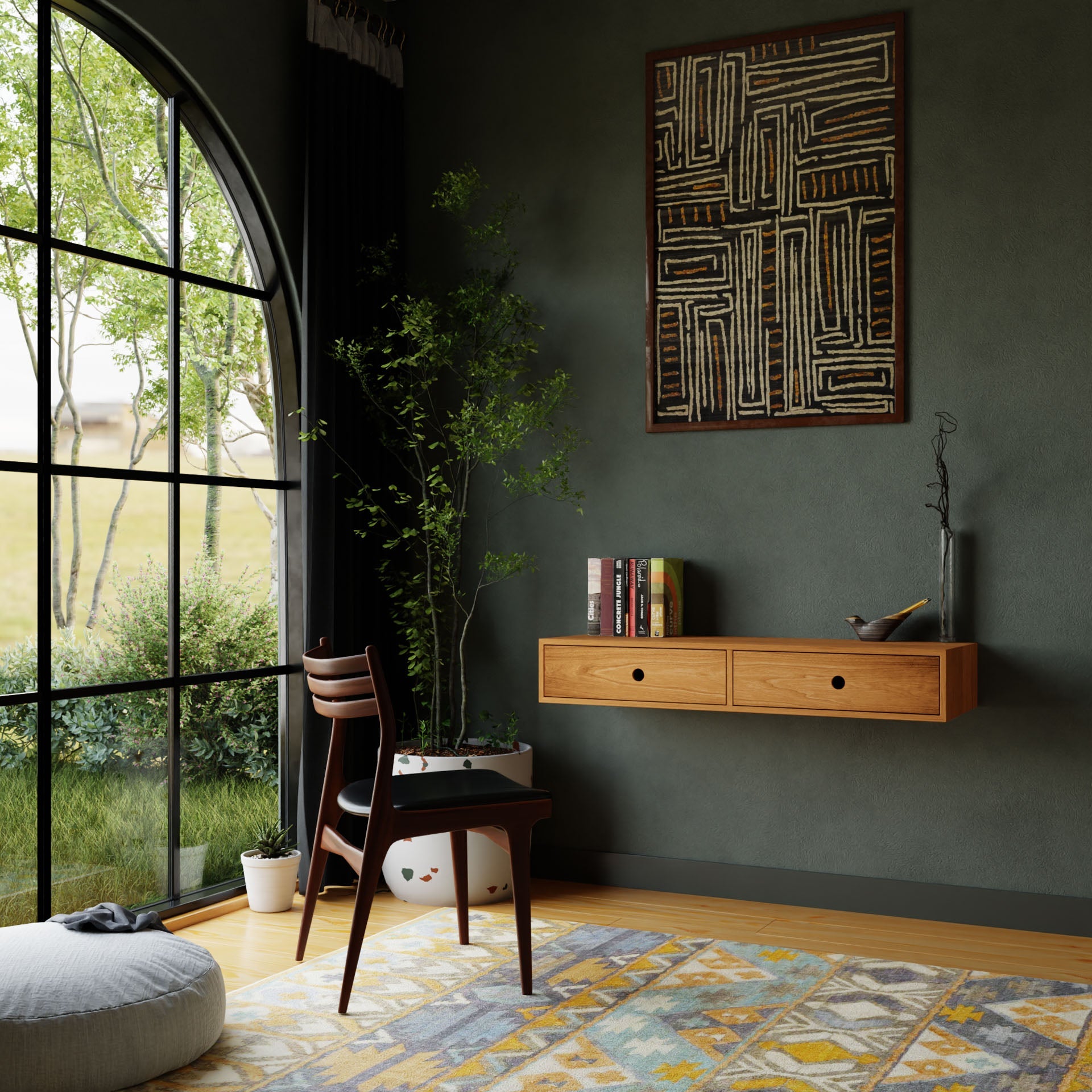 A cozy room features a large arched window with green views, a handmade wooden chair, pouf, and Krovel's Floating Desk in Cherry. Abstract art decorates the dark green wall. The floor has a vibrant geometric rug and a small potted plant.