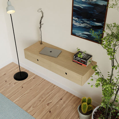 A minimalist room features a handmade Krovel Floating Desk in Maple, holding a laptop and books. A tall floor lamp stands nearby. A potted plant and cacti adorn the wooden floor, while a framed abstract painting hangs on the white wall.