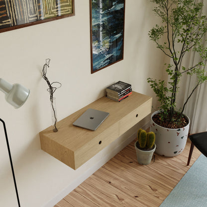 A minimalist room showcases a Krovel Floating Desk in Maple, holding a closed laptop and books. Leafy plants and a cactus bring nature beside it, while two art pieces hang above alongside a white lamp. Light wood flooring enhances the handmade aesthetic.