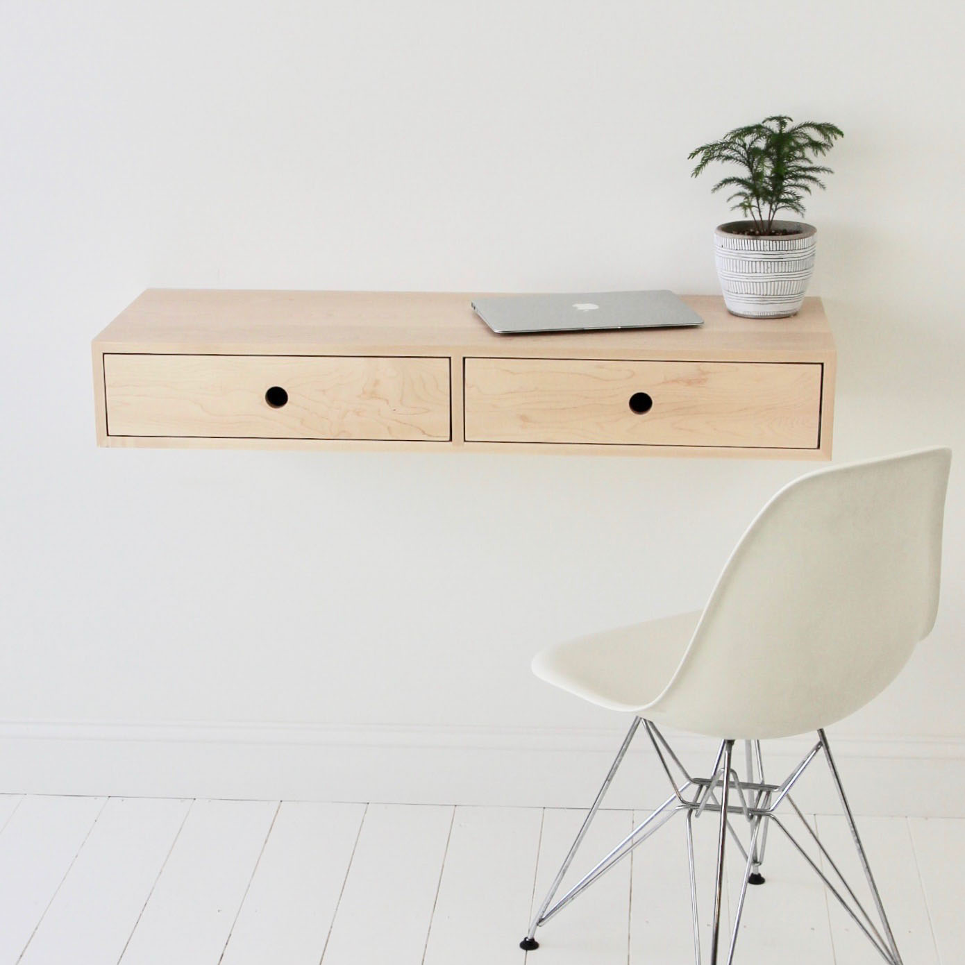 A minimalist workspace showcases a sleek floating desk in Maple by Krovel, featuring two drawers. A closed laptop and a small potted plant sit on top. A white modern chair with metal legs rests on a white wooden floor against a plain wall, exuding elegance and simplicity.