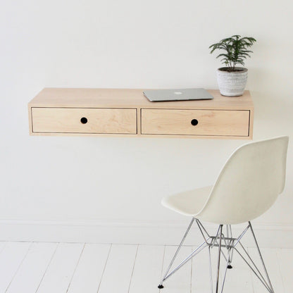A minimalist workspace showcases a sleek floating desk in Maple by Krovel, featuring two drawers. A closed laptop and a small potted plant sit on top. A white modern chair with metal legs rests on a white wooden floor against a plain wall, exuding elegance and simplicity.