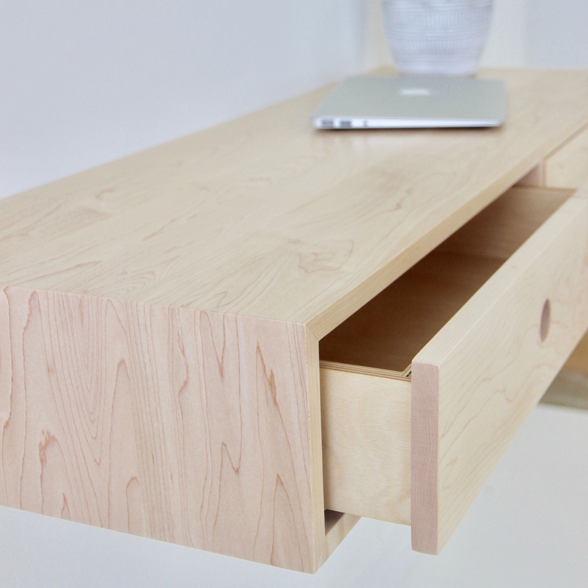 Close-up of a light, handmade wooden Floating Desk in Maple by Krovel with a partially open drawer. A silver laptop rests on this elegant surface, and a partially visible striped vase is in the background, accentuating the prominently visible wood grain.