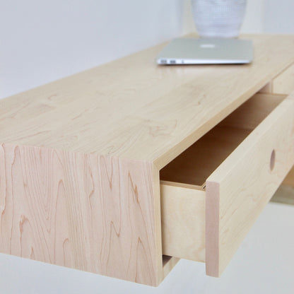 Close-up of a light, handmade wooden Floating Desk in Maple by Krovel with a partially open drawer. A silver laptop rests on this elegant surface, and a partially visible striped vase is in the background, accentuating the prominently visible wood grain.