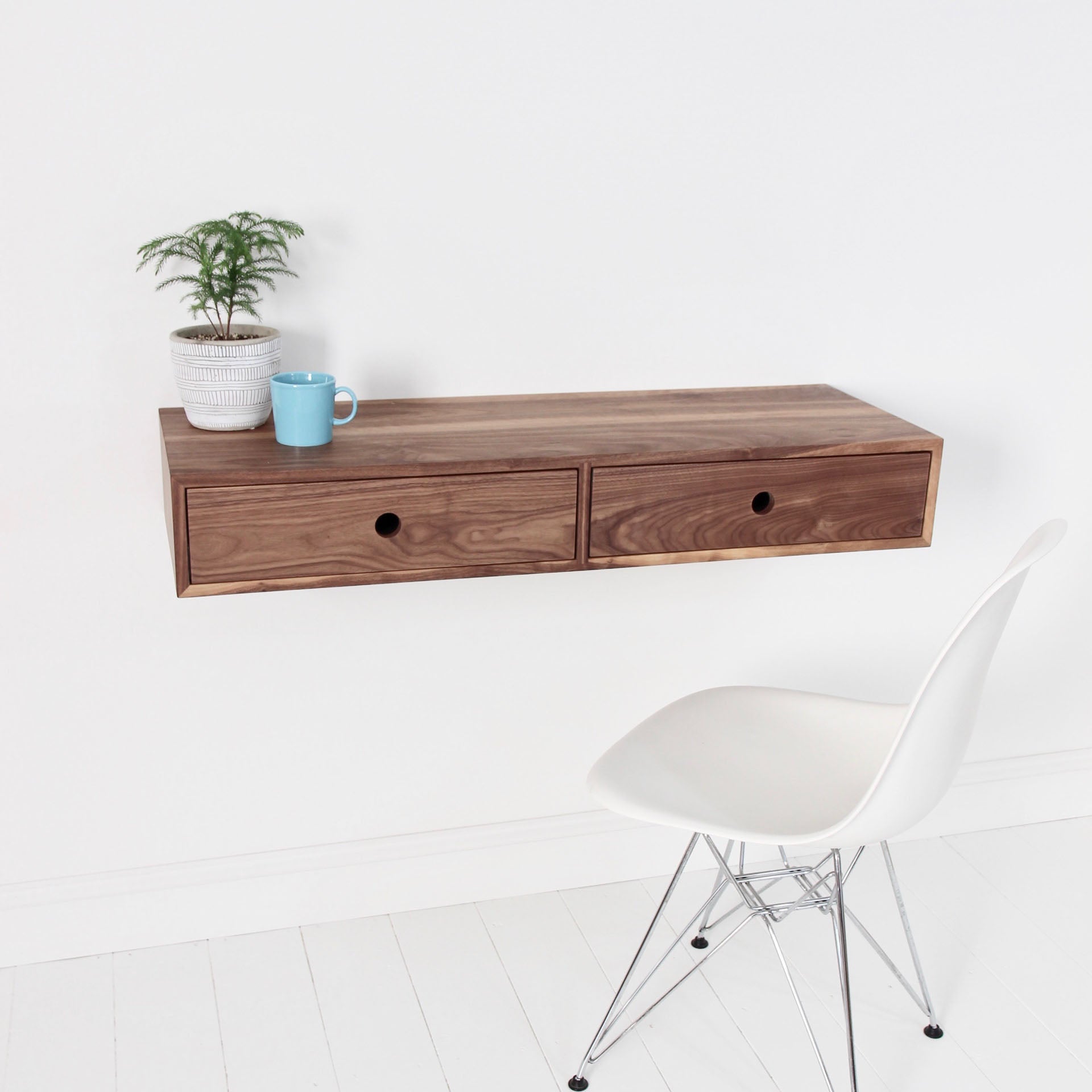 A minimalist workspace features Krovel's handmade Floating Desk in American Black Walnut with two drawers. Adorning the top are a potted plant and a blue mug. Below is a sleek, modern white chair with metal legs on the pristine white wooden floor.