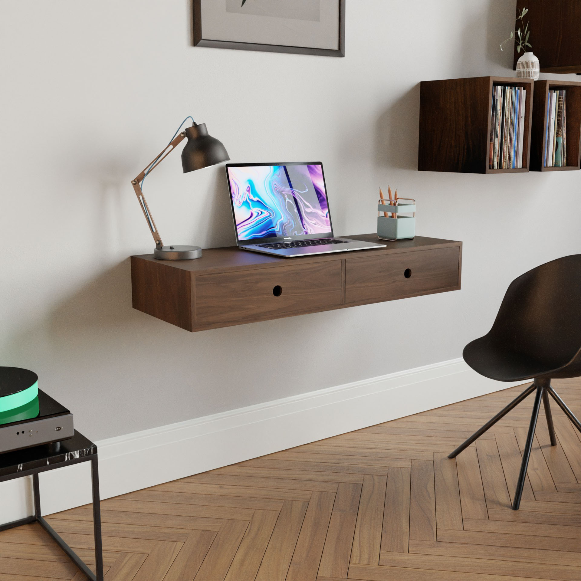 A minimalist workspace includes Krovel's Floating Desk in Walnut, holding a laptop, lamp, pen holder, and small plant. Nearby is a sleek black chair, and above the herringbone-patterned floor are wall-mounted shelves with books and decor.