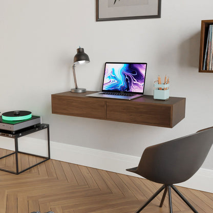 A minimalist workspace features the Krovel Floating Desk in Walnut, displaying a laptop with a vibrant abstract screen. A handmade lamp and pencil-filled cup sit on it. Nearby is a black chair, while a record player rests on a corner stand atop the wooden floor.