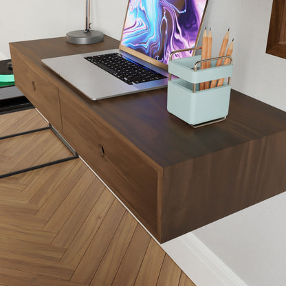 A Krovel Floating Desk in Walnut holds an open laptop with a colorful display, accompanied by a silver lamp and a light blue pencil holder containing three pencils. The herringbone wood pattern floor enhances the scene.