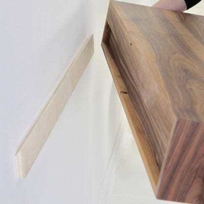 Someone is installing a Krovel Floating Desk in Walnut onto a wall-mounted bracket. The rich American Black Walnut desk, with its dark wood grain, beautifully contrasts the lighter bracket and elegantly stands out against the white-painted wall.