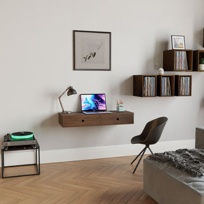 A modern home office features a Krovel Floating Desk in Walnut, adorned with a laptop, lamp, and pen holder. A framed picture hangs above, while shelves house records and books. The space is completed with a chair, record player, and sofa on wooden flooring for a cozy ambiance.