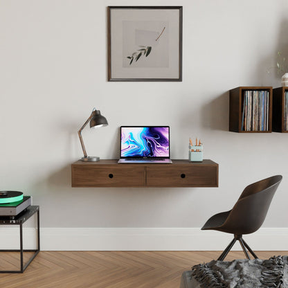 In a modern home office, a Krovel Floating Desk in Walnut holds a laptop showing an abstract design. A silver lamp and pencils in a holder are functional accents. A framed artwork hangs above, with shelves of vinyl records on the right. A black chair sits nearby.