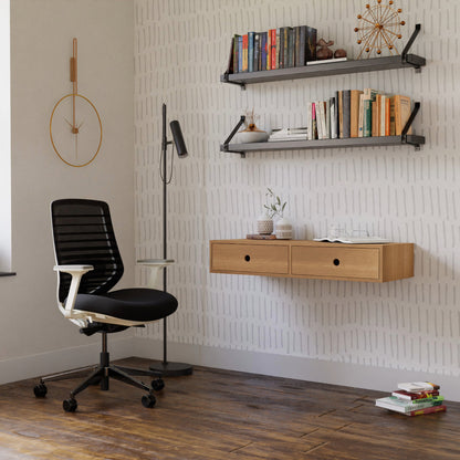 A modern home office is equipped with a sleek black chair and Krovel's hand-finished, wall-mounted Floating Desk in White Oak. Floating shelves hold books and decor, such as a geometric sculpture and potted plant. A floor lamp stands beside the desk against a patterned wall.