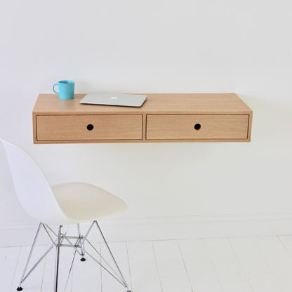 A minimalist setup includes a Krovel Floating Desk in White Oak, wall-mounted with two drawers and supporting a closed laptop and blue mug. Nearby, a white modern chair with wooden legs sits on the white floor against a plain white wall.