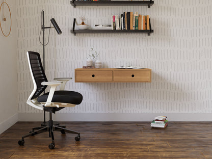 A modern office features a hand-finished black and white ergonomic chair on a wooden floor. A wall-mounted Krovel floating desk in White Oak holds a vase and books. Above, two shelves showcase books and decor against patterned wallpaper, while a tall black lamp stands nearby.