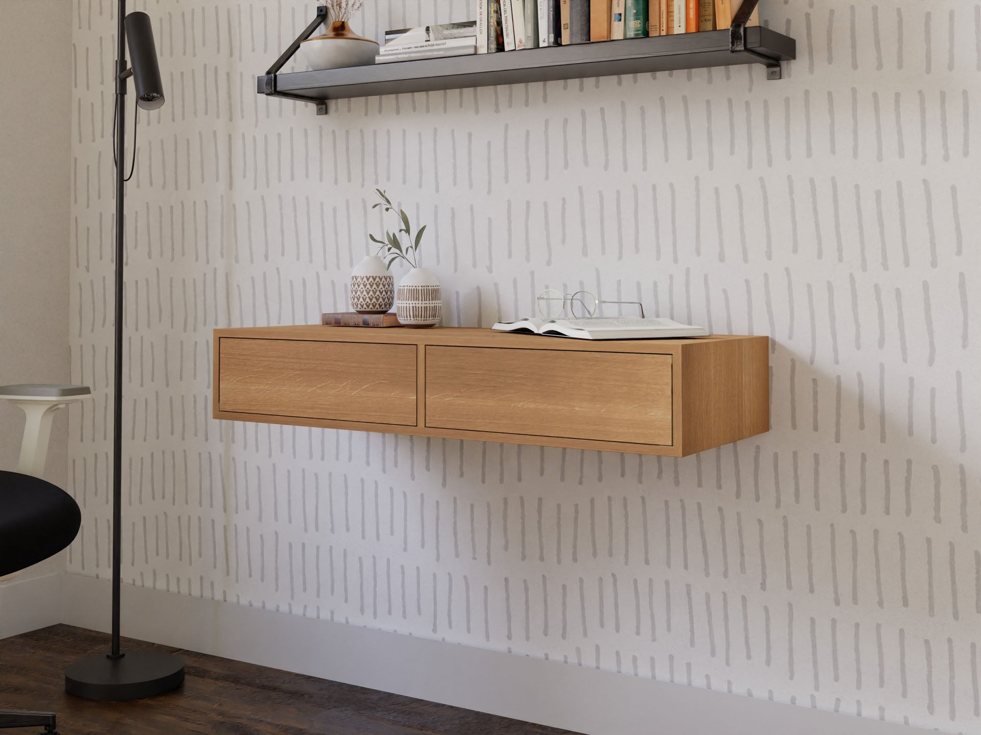 In a minimalist room, a Krovel Floating Desk in White Oak is mounted against a patterned wall. The desk displays small potted plants and an open book. Above, a black shelf carries books, while nearby, a black floor lamp stands on dark wooden flooring.