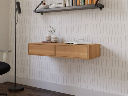 In a minimalist room, a Krovel Floating Desk in White Oak is mounted against a patterned wall. The desk displays small potted plants and an open book. Above, a black shelf carries books, while nearby, a black floor lamp stands on dark wooden flooring.