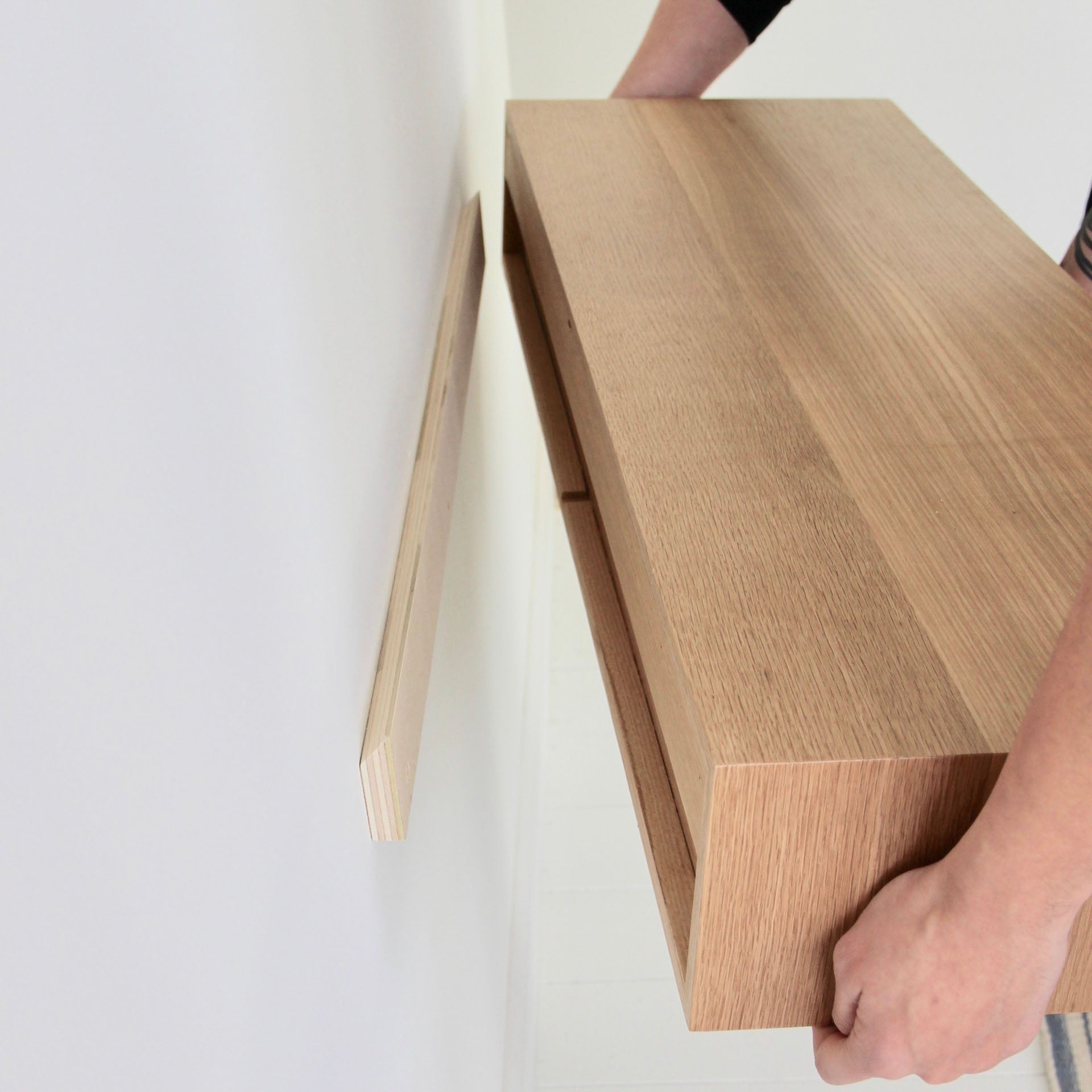 A person is mounting a hand-finished Floating Desk in White Oak from Krovel on the wall. The desk, with a light and elegant finish, is being aligned with a narrow wooden bracket already attached to the wall. Careful hands are visible holding the desk in place.