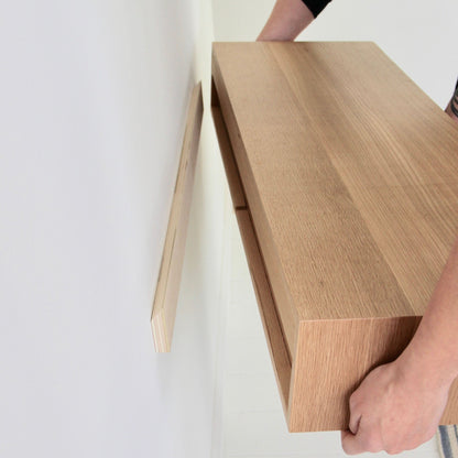 A person is mounting a hand-finished Floating Desk in White Oak from Krovel on the wall. The desk, with a light and elegant finish, is being aligned with a narrow wooden bracket already attached to the wall. Careful hands are visible holding the desk in place.