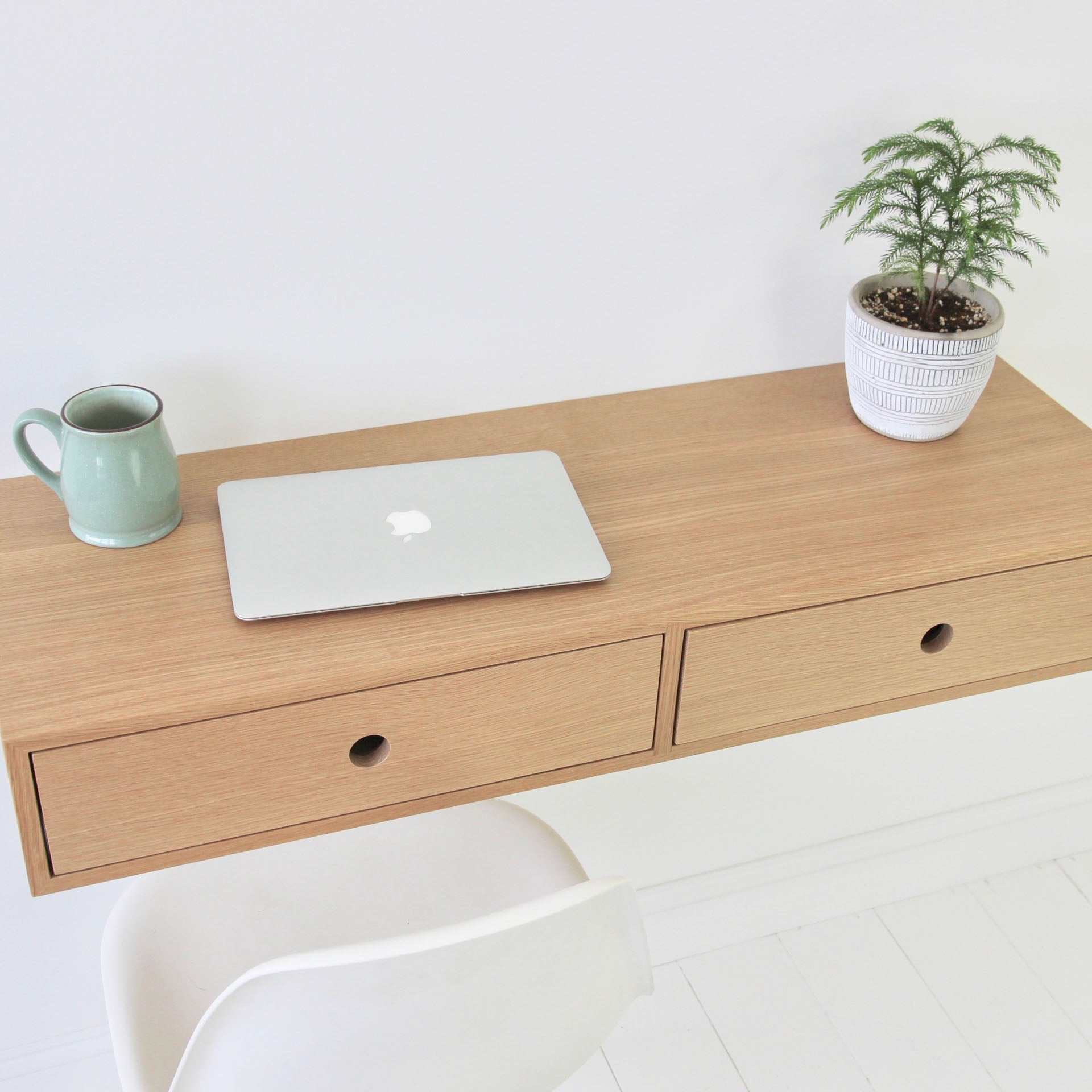 A minimalist workspace features the Krovel Floating Desk in White Oak, hand-finished with two drawers. On the desk are a closed silver laptop, a green mug, and a small potted plant. A white chair peeks into view against the serene white wall background.
