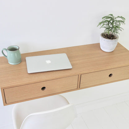 A minimalist workspace features the Krovel Floating Desk in White Oak, hand-finished with two drawers. On the desk are a closed silver laptop, a green mug, and a small potted plant. A white chair peeks into view against the serene white wall background.
