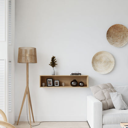 A minimalist living room with a light sofa, wooden tripod floor lamp, framed photos and decor on the Krovel Floating Display Case in Maple, and two woven wall baskets on a white wall.
